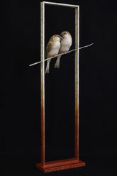Sculpture of two birds perched on a branch inside a tall, narrow frame against a black background.