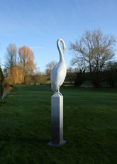 White swan sculpture on a pedestal in a park with trees and water in the background