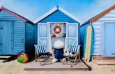 Toy character sitting on a beach chair in front of colorful beach huts with a surfboard and ball.