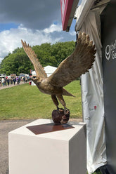 Bronze sculpture of a falcon on a pedestal outdoors with people and tents in the background.