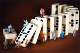 A set of large off-white dominoes arranged on a dark brown surface, with several miniature human figures pushing, balancing, and standing on the dominoes. The scene is photographed in a studio setup with no visible brand markings.