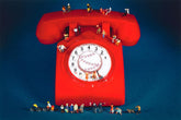 A surreal studio photograph of a giant red rotary telephone on a dark blue background, with tiny human figurines standing on and around it.
