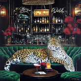 A large leopard lies stretched across a green velvet bar sofa in a luxurious bar interior, with a cocktail on a table in front and shelves of bottles behind it.