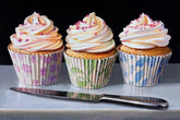 Three cupcakes with colorful frosting and decorative paper cups on a reflective surface with a knife.
