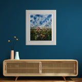 Framed artwork of a field of flowers with a blue sky on blue wall above wooden sideboard.
