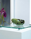 Decorative green leaf containing a green toad on a glass shelf with a purple flower framed artwork in the background