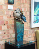 Bronze sculpture of four puffins on a pedestal in front of a brick wall with framed artwork.