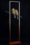 Sculpture of two birds perched on a branch inside a tall, narrow frame against a black background.