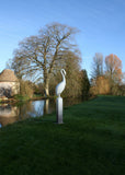 White bird sculpture on a pedestal in a park with trees and a pond in the background.