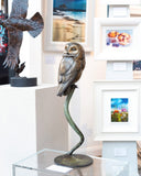 Bronze owl sculpture on a pedestal in an art gallery setting with framed pictures on the wall.