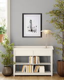 White bookshelf with books, framed picture, and lamp against a gray wall with plants.