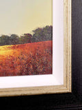 Close-up of a framed landscape painting showing a textured orange and red field with dark trees under a pale sky, with a white mat and ornate gold-toned frame.