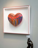 Colorful heart-shaped artwork in a white frame on a wall, with a sculpture of a hand below.