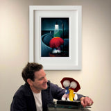Man holding a small figurine with a red umbrella next to a framed artwork on a wall.