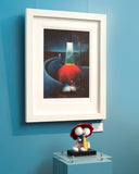 Framed artwork of a character with a red hat on a blue wall, accompanied by a small sculpture below.