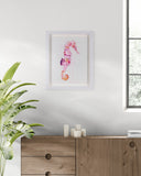 Framed artwork of a pink seahorse on a white wall above a wooden cabinet with candles.