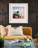 A framed art print featuring a whimsical illustration of dogs on a beach in a mint green fiat is displayed on a dark wood-paneled wall above a mustard yellow sofa with decorative pillows. The scene includes a blue wooden table with an open magazine, a small green car sculpture with dog characters, and potted plants.