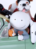 A close-up of a stylized figurine or collectible set featuring cartoon dog and cat characters sitting in a light green car. The central figure is a large white character with a simple black smiley face, surrounded by smaller dog and cat figures in white, black, orange, brown, and red.