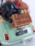 A close-up of a decorative figurine showing a black dog sitting in the back of a mint green car, holding a brown wicker picnic basket. The figurine includes a tan jacket and a black license plate reading 'DH 12' on a white background.