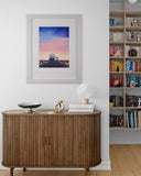A framed art print featuring a colorful sunset sky with a silhouette of a person, displayed on a white wall above a wooden sideboard in a styled room with a bookshelf.