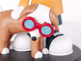 A close-up of a figurine featuring a brown and white dog wearing red-rimmed glasses with light blue lenses, standing in front of a person in an orange top and dark pants on a black circular base.