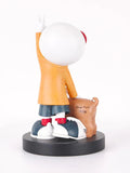 A stylized figurine of a person with a large white head, wearing an orange top, blue shorts, and white sneakers with red accents, standing on a black circular base and holding a brown dog. The figurine is photographed against a clean white background.
