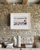 A framed art print depicting a scenic coastal landscape with a figure and small dog, displayed on a rustic stone wall above a styled coffee table in a living room setting.