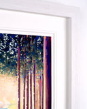A framed art print featuring an impressionistic painting of tall trees with blue, purple, and warm golden tones, shown in a white frame against a white wall.