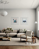 A styled living room interior featuring a light beige sectional sofa, a matching armchair with wooden arms, a low wooden coffee table with a vase of pink flowers, and two framed art prints hanging on a light gray wall.