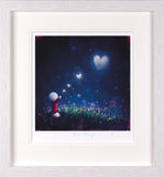 A framed art print featuring a whimsical illustration of a small figure in a field under a starry night sky with glowing heart-shaped stars, presented in a white mat and light wood frame.