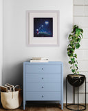 A framed art print depicting a whimsical fairy in a glowing field under a heart-shaped moon, styled in a bright room above a light blue three-drawer dresser.