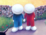 A stylized painting of two smiling stick-figure-like characters standing hand in hand on a path, one in a blue outfit and one in a red outfit, with a stone wall and grassy field behind them.