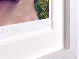 Close-up of a framed art print showing a white mat border, part of a colorful painting with green foliage and a pinkish-purple surface, and a small handwritten signature.