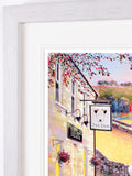 Framed art print featuring a colorful painting of a quaint village pub exterior with a hanging 'The Dog' sign, set in a white frame with a white mat.