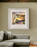 A framed art print depicting a whimsical village street scene with a couple and a black dog outside a country pub, displayed on a beige wall above a grey sectional sofa.