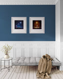 A styled interior scene with a blue accent wall, white wainscoting, two framed abstract prints, a light gray tufted bench, a beige throw blanket, a small round side table with a vase of branches, and light wood flooring.