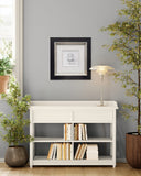 White bookshelf with books, lamp, and framed picture against a gray wall with plants.