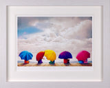 A framed art print depicting five colorful umbrellas on a wooden deck under a dramatic cloudy sky, presented in a white frame with a white mat.