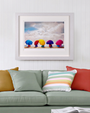 A framed art print depicting colorful beach umbrellas under a cloudy sky, displayed on a white paneled wall above a green sofa with decorative pillows.