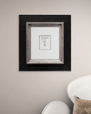 Framed black and white artwork on a wall with a chair and pillow in the foreground.