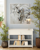 A framed abstract painting of a horse head in white, gray, and black tones, displayed on a light blue wall above a white console table with books and a lamp, surrounded by potted plants.