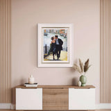 Framed artwork of a couple by a canal on a wall above a wooden and white cabinet.
