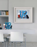 A framed art print featuring a colorful 'HOME' design with a red and white figure, a black dog, and blue lettering, displayed on a light gray wall above a white dining table with chairs.