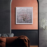 Framed artwork of a cherry blossom tree on a wall with a sofa and plant in the foreground.