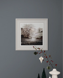 Framed black and white landscape print on a gray wall with decorative branches.