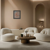 A modern living room scene with three rounded off-white upholstered chairs arranged around a low round wooden coffee table. A beige wall, arched alcove, shelving, and a framed artwork are visible in the background.