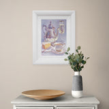 Framed artwork of a teapot and cup on a wall above a table with a vase and plate.