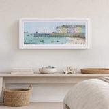 A framed horizontal art print depicting a colorful coastal harbor scene with pastel buildings, boats, and a pier, displayed on a light wall above a shelf with decorative items.