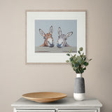 Framed artwork of two donkeys on a wall above a table with a vase and plate.