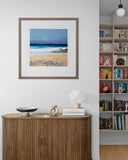 Framed beach scene above a wooden sideboard with decor items in a room.